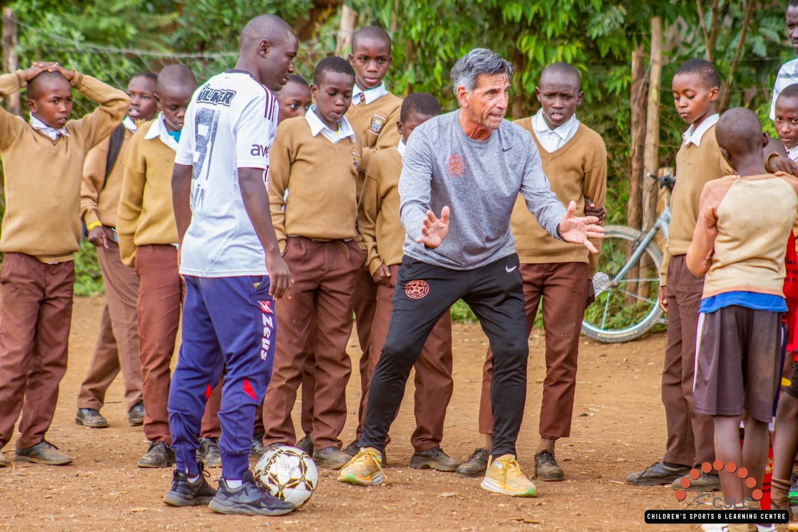A coach demonstrating a drill to a circle of students on a dirt field